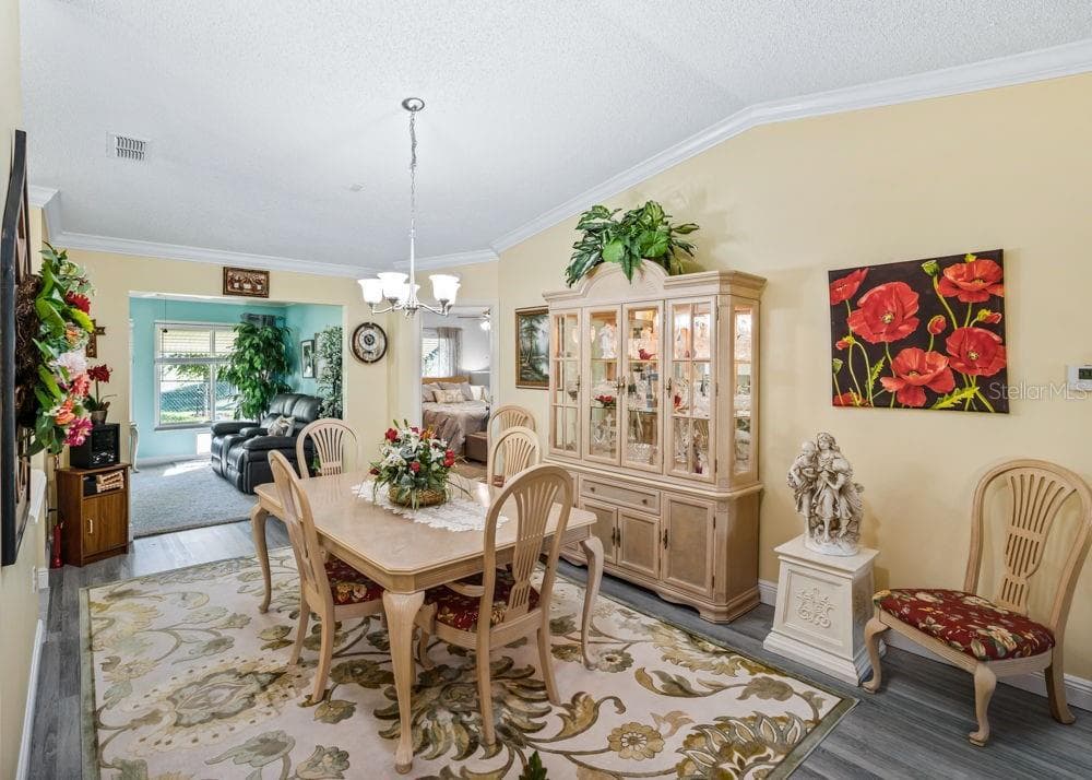 Inviting dining area featuring wood-look vinyl flooring, crown molding, and a vaulted ceiling with a chandelier. The open layout flows into a sunlit sitting area, creating a welcoming space for everyday meals and entertaining. - 17909 SE 99TH AVE, SUMMERFIELD