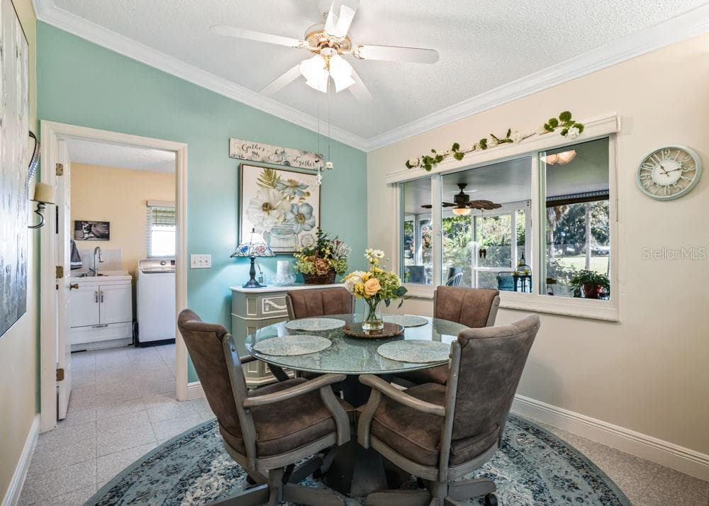 Charming breakfast nook featuring a soft aqua accent wall, crown molding, and a ceiling fan. A large interior window overlooks the enclosed lanai and greenery beyond, while the adjacent laundry room adds everyday convenience. - 17909 SE 99TH AVE, SUMMERFIELD