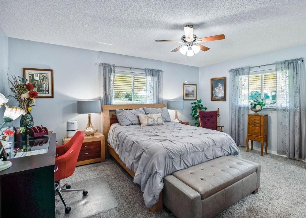 Spacious primary bedroom featuring soft blue walls, carpeting, and a ceiling fan with lighting. Two windows welcome natural light, while the generous layout offers room for a desk or cozy reading area. - 17909 SE 99TH AVE, SUMMERFIELD