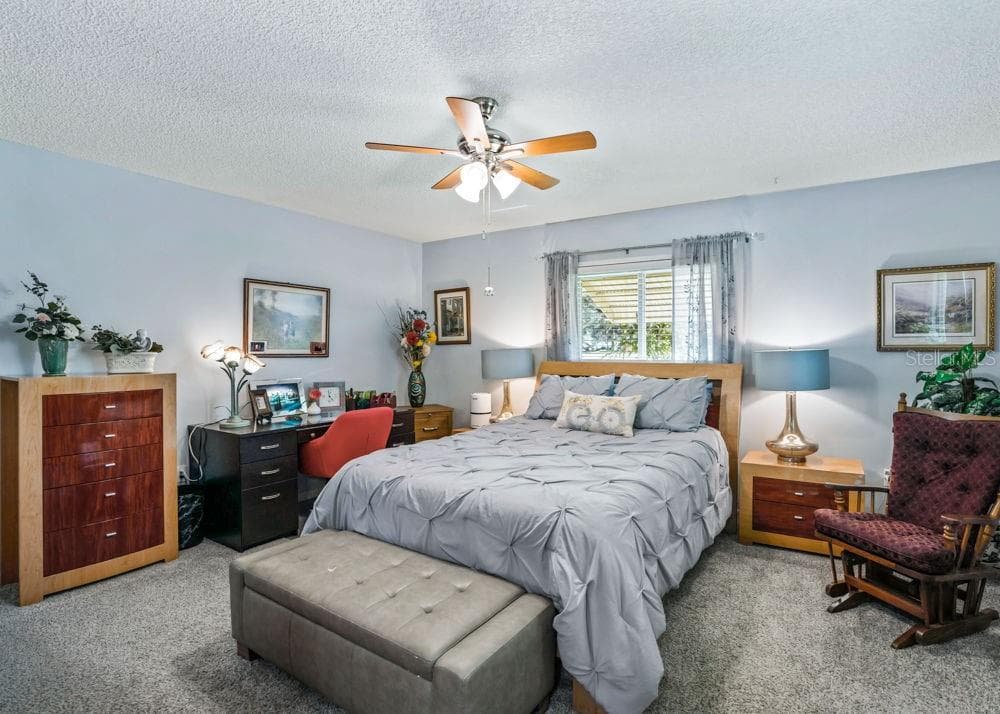 The spacious primary bedroom offers soft blue walls, comfortable carpeting, and a ceiling fan with lighting. Natural light filters through the window, creating a relaxing retreat with space for a desk or cozy sitting area. - 17909 SE 99TH AVE, SUMMERFIELD