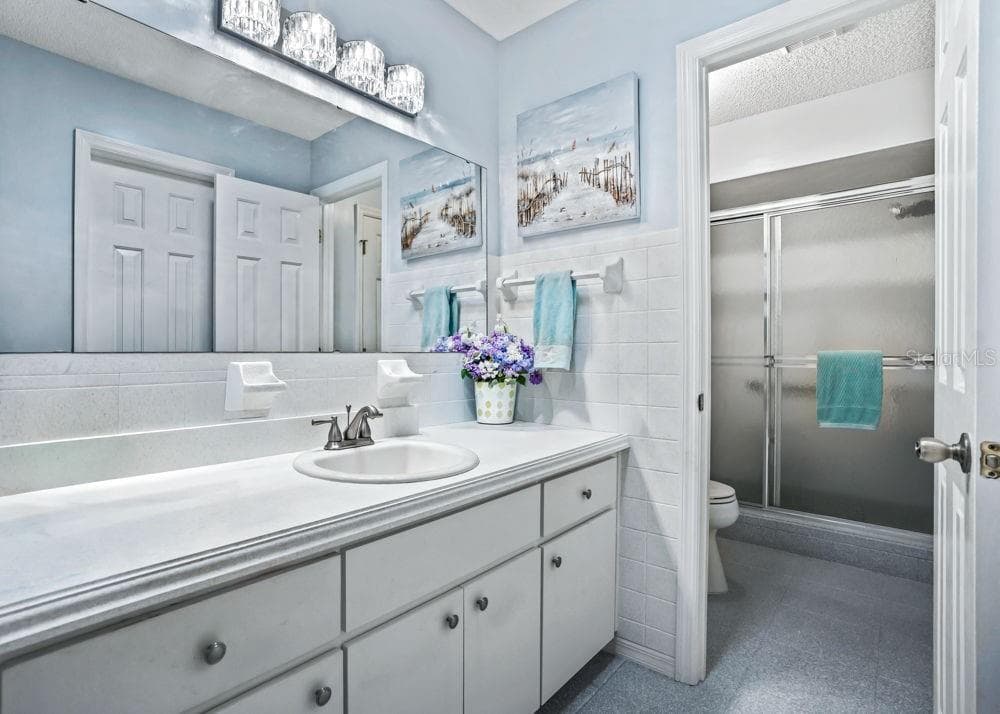 The primary bathroom features a spacious vanity with generous counter space, ample cabinet and drawer storage, and a large mirror with decorative lighting. Soft blue walls and white tile accents create a bright, relaxing setting, while a separate area contains the toilet and walk-in shower with sliding glass doors. - 17909 SE 99TH AVE, SUMMERFIELD