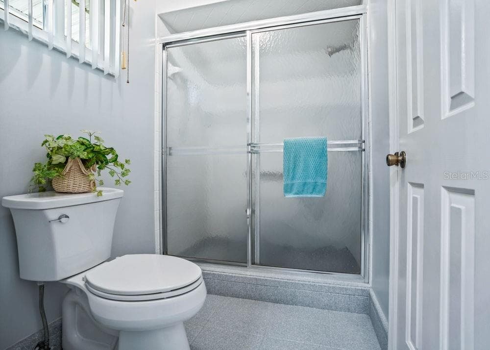 The primary bathroom features a spacious walk-in shower with frosted sliding glass doors, neutral flooring, and soft blue walls. A high window brings natural light into the space while maintaining privacy. - 17909 SE 99TH AVE, SUMMERFIELD