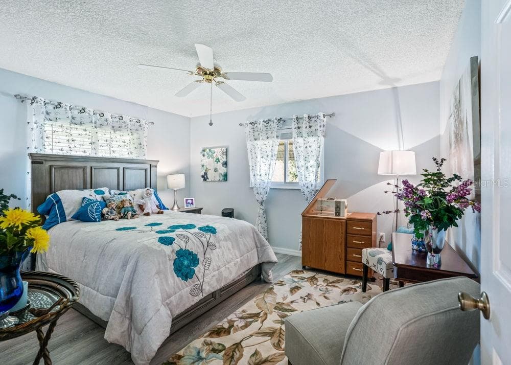 The second bedroom features soft blue walls, wood-look flooring, and two windows that fill the room with natural light. A ceiling fan adds comfort, while the layout provides space for a bed, desk, and additional seating. - 17909 SE 99TH AVE, SUMMERFIELD