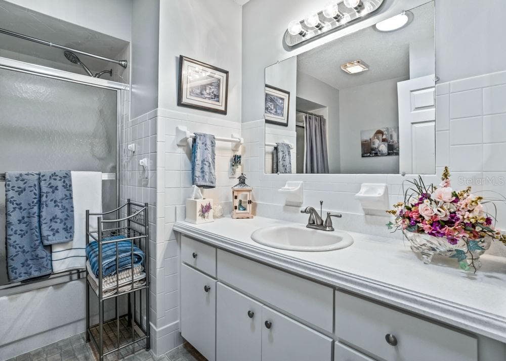 The second bathroom features a spacious white vanity with ample counter space and storage, a large mirror, and bright vanity lighting. White tile accents and a tub/shower combination with frosted sliding glass doors complete the space. - 17909 SE 99TH AVE, SUMMERFIELD