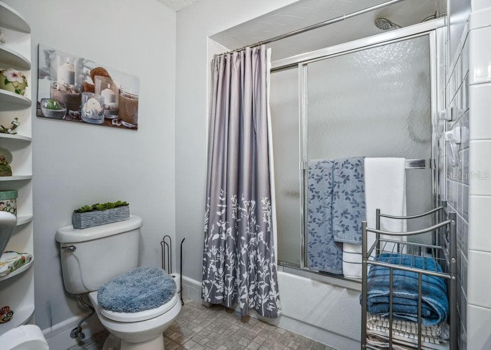 Another view of the second bathroom highlights the tub/shower combination with frosted sliding glass doors and white tile surround. Soft neutral walls, patterned flooring, and built-in corner shelving complete the space. - 17909 SE 99TH AVE, SUMMERFIELD