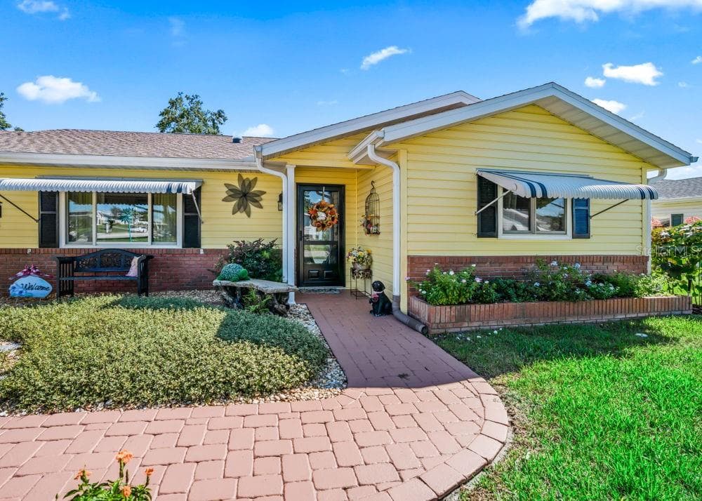 A curved brick-paver walkway leads to the welcoming covered front entry, framed by freshly painted cheerful yellow siding, brick accents, and landscaped garden beds. Striped window awnings, dark shutters, and a glass storm door add to the home’s inviting curb appeal. - 17909 SE 99TH AVE, SUMMERFIELD