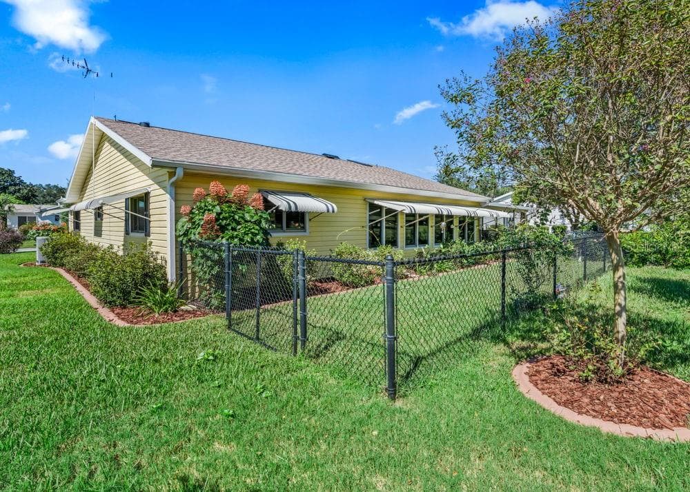 The fenced backyard features lush green lawn, flowering plants, and mature landscaping, with a gated entrance for convenient access. A brick-paver walkway borders the home, while striped window awnings and cheerful yellow siding add charm to the outdoor setting. - 17909 SE 99TH AVE, SUMMERFIELD