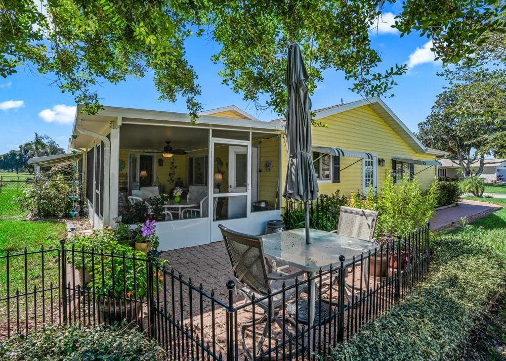 The inviting rear brick-paver patio is surrounded by lush landscaping, colorful flowers, and large shade trees, creating a peaceful outdoor retreat. Decorative black fencing and direct access to the screened lanai make this a charming spot for outdoor dining or relaxing. - 17909 SE 99TH AVE, SUMMERFIELD