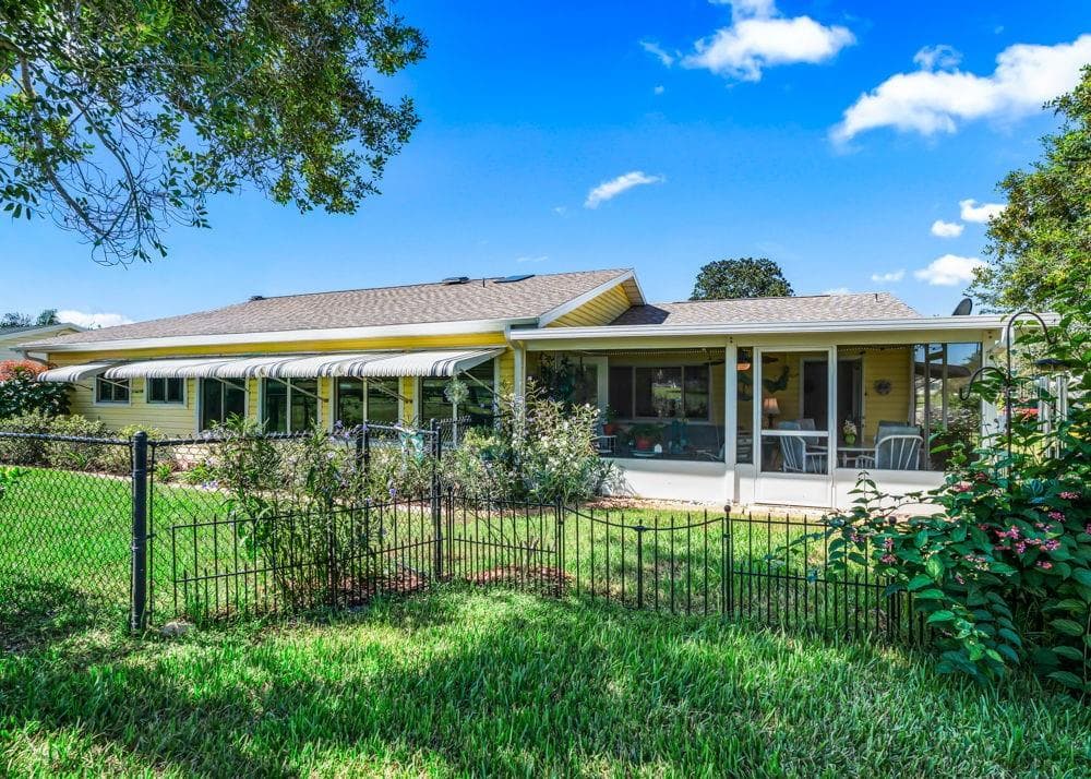 This rear exterior view showcases the spacious screened lanai, window-lined Florida room, and striped awnings against cheerful yellow siding. A fenced backyard with lush lawn, flowering plants, and mature shade trees creates an inviting outdoor setting. - 17909 SE 99TH AVE, SUMMERFIELD