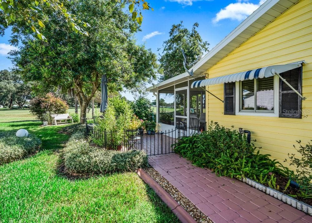 A brick-paver walkway leads around the house to the gated back patio and screened lanai, bordered by lush landscaping and mature shade trees. Cheerful yellow siding, striped window awnings, and views of The Links golf course add to the inviting outdoor setting. - 17909 SE 99TH AVE, SUMMERFIELD