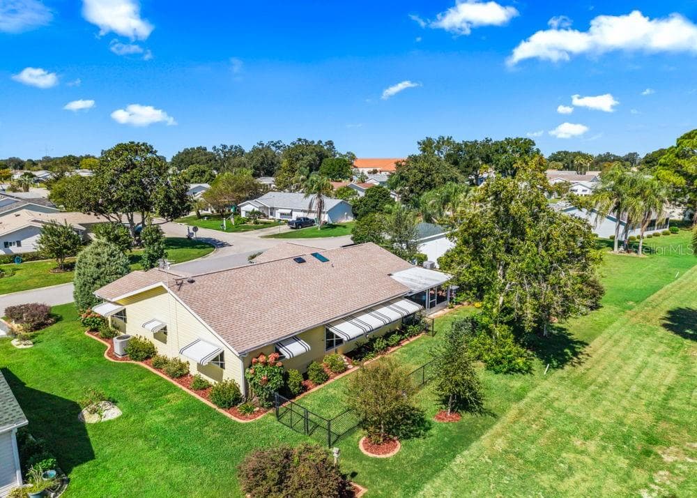 This aerial rear view highlights the home’s golf course setting, fenced backyard, and mature shade trees. Landscaped garden beds, striped window awnings, and a screened lanai complement the cheerful yellow exterior, creating an inviting space to enjoy Florida living. - 17909 SE 99TH AVE, SUMMERFIELD