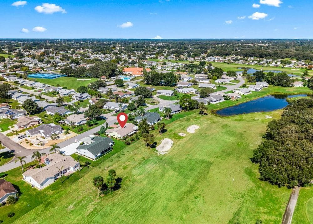 This aerial view highlights the home’s location directly along The Links golf course, marked by the red pin, with expansive fairways and a nearby pond. The wider view showcases the surrounding neighborhood, mature trees, and scenic green spaces. - 17909 SE 99TH AVE, SUMMERFIELD