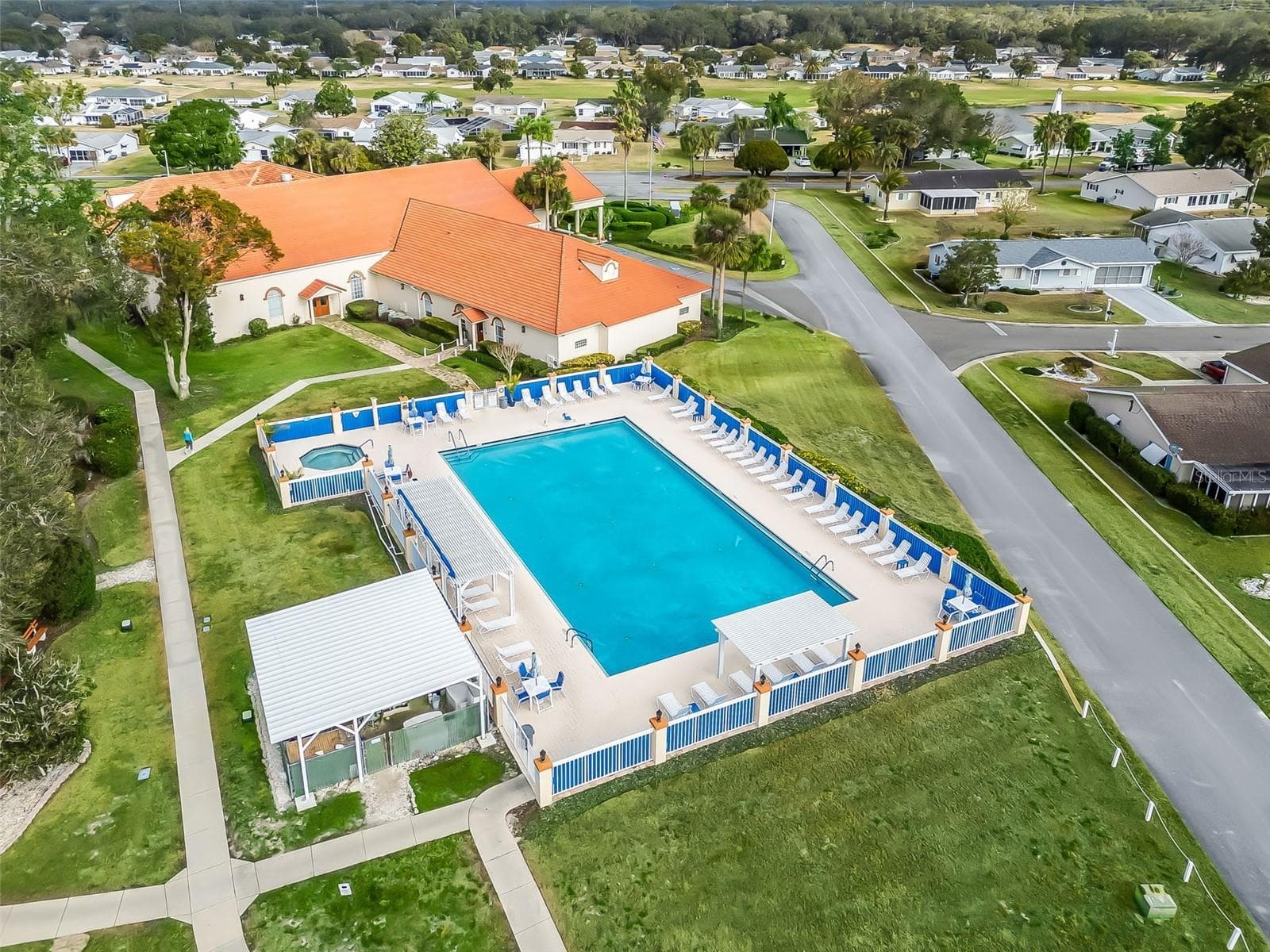 This aerial view showcases Spruce Creek South’s community swimming pool and spa, surrounded by a spacious sun deck with lounge chairs and covered seating areas. The adjacent clubhouse and landscaped grounds create an inviting setting for relaxation and socializing. - 17909 SE 99TH AVE, SUMMERFIELD