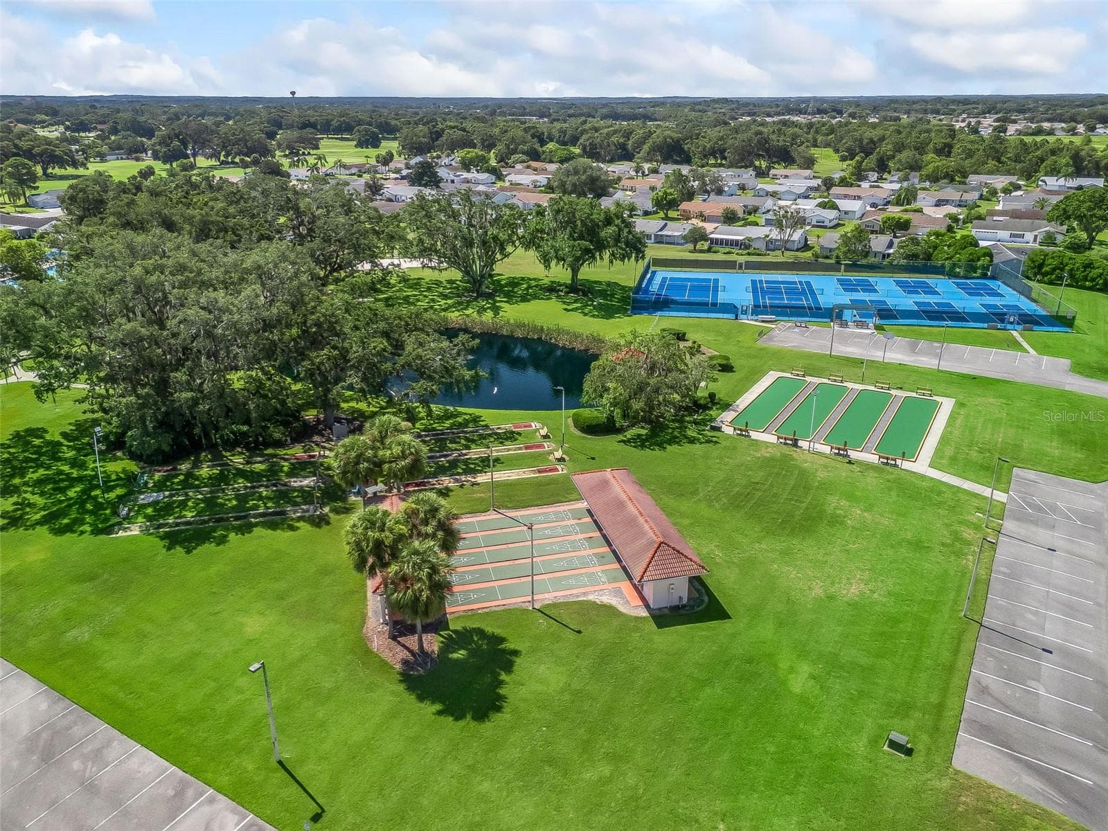 This aerial view highlights Spruce Creek South’s outdoor recreation area, featuring tennis and pickleball courts, shuffleboard courts, and a scenic pond. Expansive lawns, mature shade trees, and nearby seating create an inviting setting for recreation and relaxation. - 17909 SE 99TH AVE, SUMMERFIELD