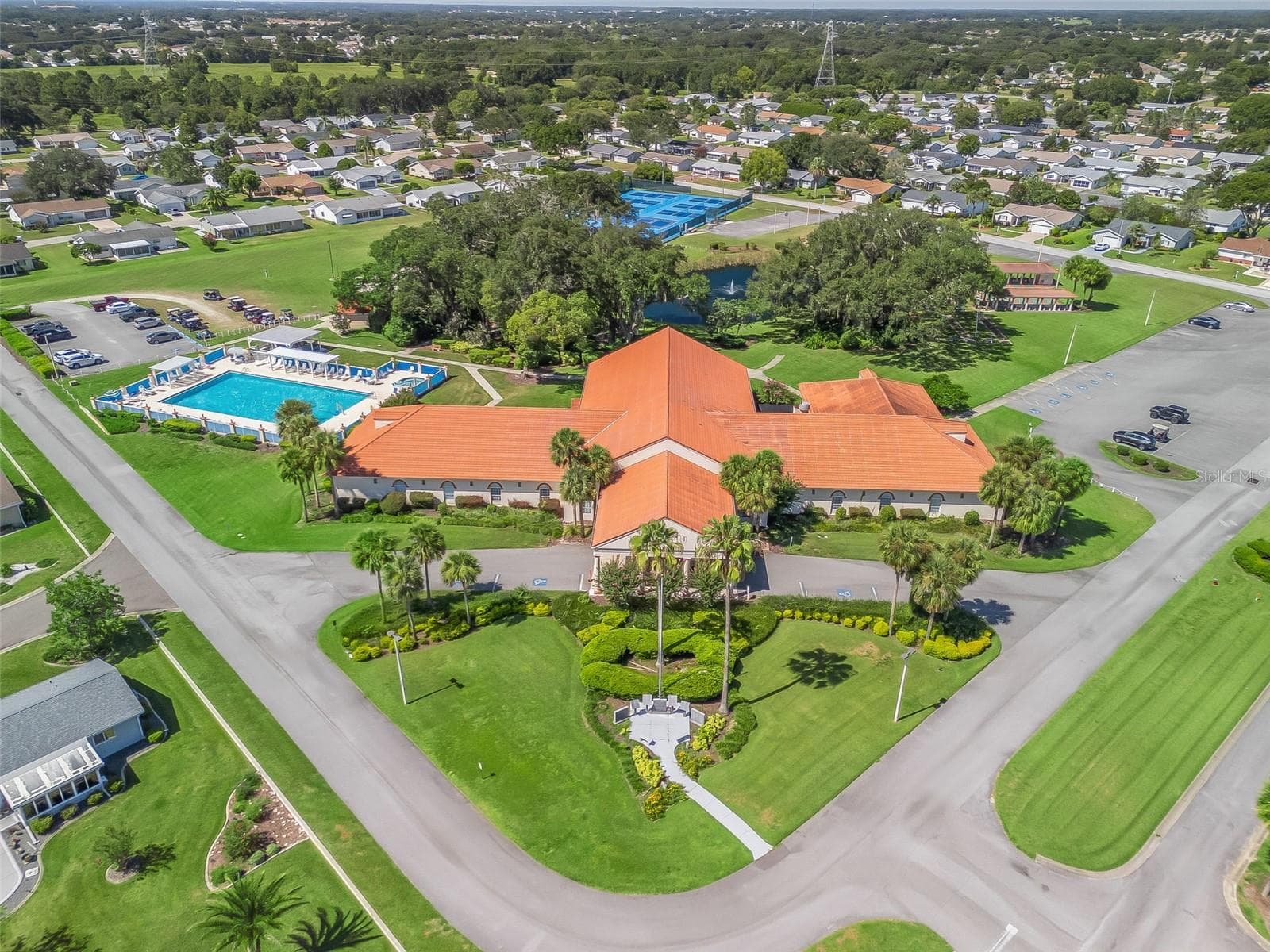 This aerial view showcases Spruce Creek South’s clubhouse, featuring a distinctive terracotta-colored roof, palm-lined entrance, and beautifully landscaped grounds. The surrounding recreation area includes a swimming pool, tennis and pickleball courts, and a scenic pond with a fountain. - 17909 SE 99TH AVE, SUMMERFIELD