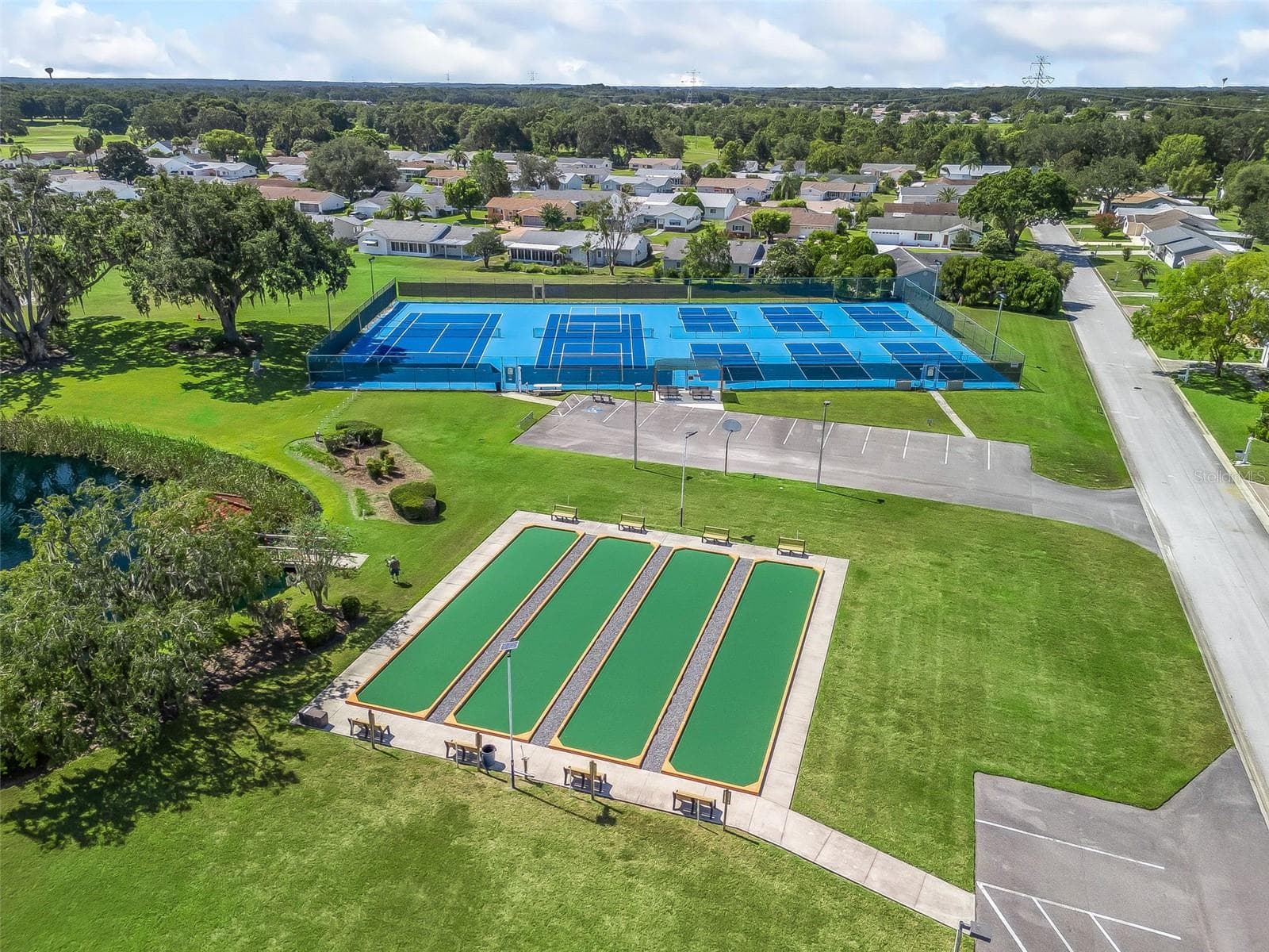 This aerial view showcases Spruce Creek South’s bocce ball courts with nearby benches, along with tennis and pickleball courts. Lush lawns, mature trees, and a scenic pond surround the recreation area, creating an inviting setting for outdoor activities. - 17909 SE 99TH AVE, SUMMERFIELD