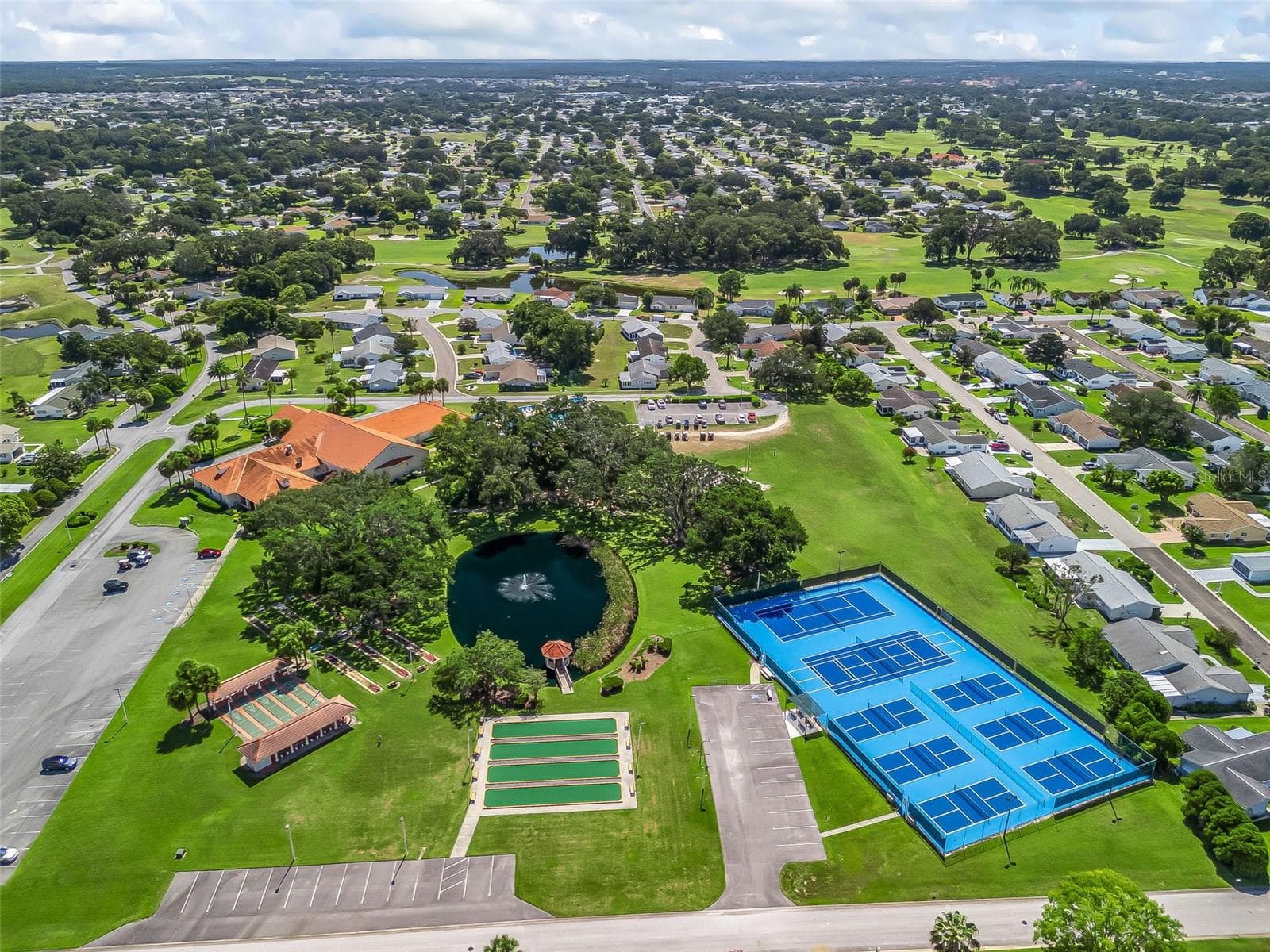 This sweeping aerial view showcases Spruce Creek South’s recreation area, featuring tennis and pickleball courts, bocce ball courts, shuffleboard, and a scenic pond with a fountain. Mature shade trees surround the nearby clubhouse, with The Links golf course and neighborhood homes extending into the distance. - 17909 SE 99TH AVE, SUMMERFIELD