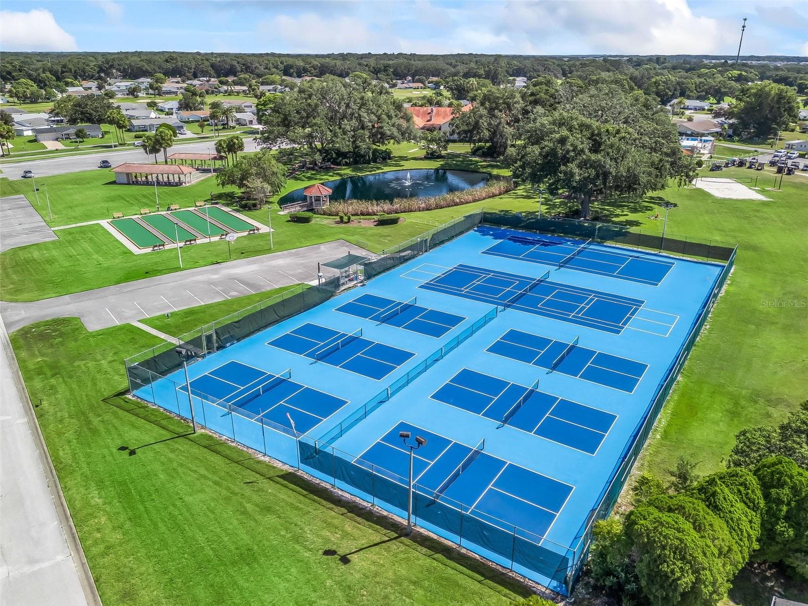 This aerial view showcases Spruce Creek South’s tennis and pickleball courts, complete with perimeter fencing and court lighting. Nearby bocce ball courts, covered seating, and a scenic pond with a fountain and gazebo enhance the inviting recreation area. - 17909 SE 99TH AVE, SUMMERFIELD