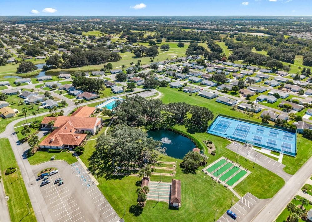 This aerial view showcases Spruce Creek South’s recreation area, featuring the clubhouse, swimming pool, tennis and pickleball courts, shuffleboard, and bocce courts. A scenic pond with a fountain, mature shade trees, and surrounding golf course views complete the inviting community setting. - 17909 SE 99TH AVE, SUMMERFIELD